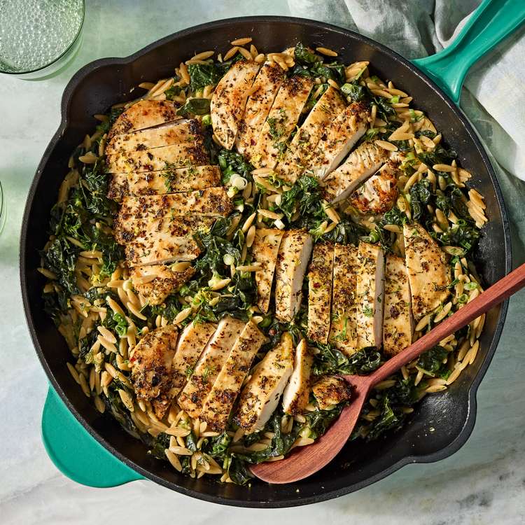 Sliced chicken cutlets nestled over creamy orzo and kale in a teal cast-iron skillet, garnished with dill