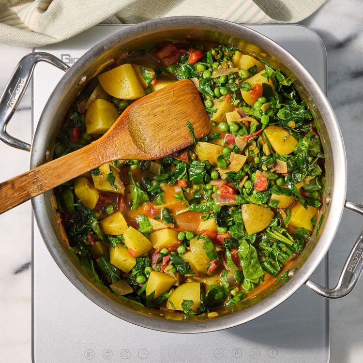Finished saag aloo matar in a stainless skillet — wilted spinach and kale, tender potatoes, peas and tomato gravy with a wooden spoon