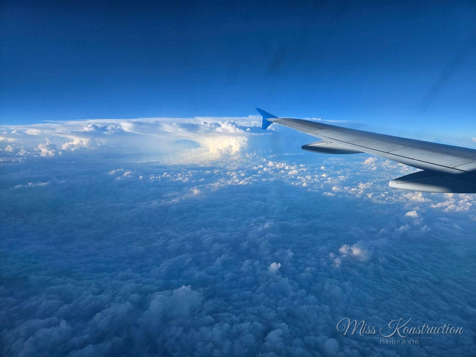 View from an airplane window — a wing cutting across a soft blanket of clouds under deep blue sky